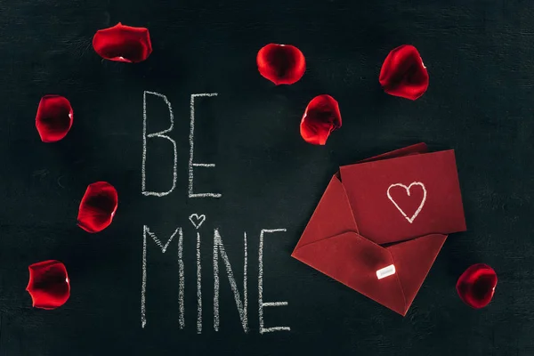 Lettering BE MINE surrounded with rose petals and red envelope on black surface — Stock Photo