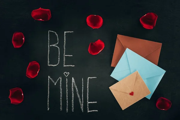 Letras BE MINE rodeado de pétalas de rosa e envelopes na superfície preta, conceito dia de São Valentim — Fotografia de Stock