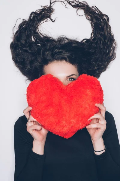 Jeune femme avec des cheveux en forme de cornes couvrant le visage avec un oreiller en forme de coeur isolé sur blanc — Photo de stock