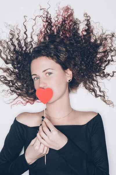Junge Frau mit lockigem Haar bedeckt Mund mit rotem Herz isoliert auf weiß — Stockfoto