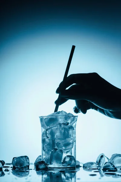 Vista recortada de la persona con paja y vidrio lleno en cubos de hielo de fusión - foto de stock