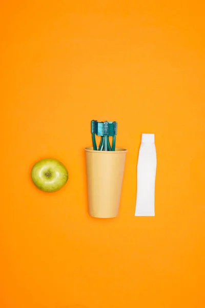 Vista superior de manzana, cepillos de dientes en taza de papel y tubo de pasta de dientes, aislado en naranja - foto de stock