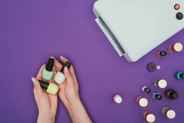 Imagen recortada de la mujer sosteniendo esmaltes de uñas en manos aisladas en púrpura - foto de stock