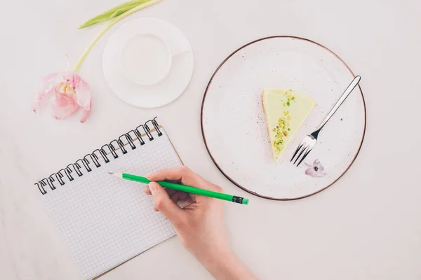 Cropped shot of woman making notes in notebook with piece of cake and cup of milk near by — Stock Photo