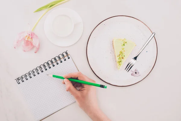 Ritagliato colpo di donna che prende appunti nel quaderno con pezzo di torta e tazza di latte nelle vicinanze — Foto stock