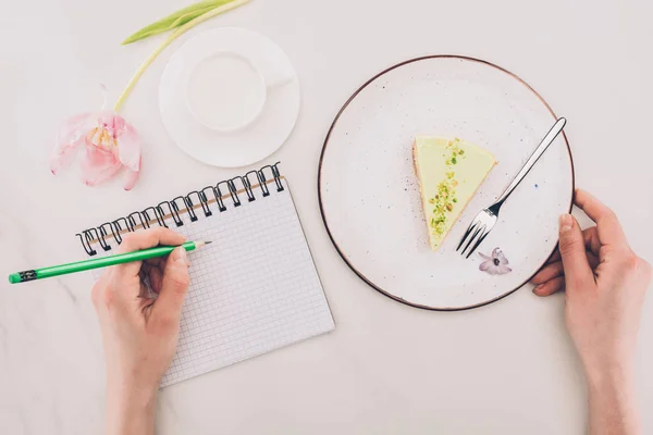Schnappschuss einer Frau, die Notizen in Notizbuch und Stück Kuchen und Tasse Milch in der Nähe macht — Stockfoto
