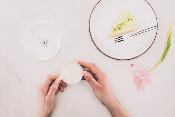Cropped shot of woman with cup of milk and piece of cake on plate on white surface — Stock Photo