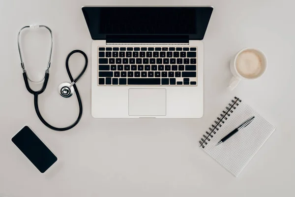 Blick von oben auf arrangiertes Stethoskop, Laptop, Notizbuch und Tasse Kaffee am Arbeitsplatz des Arztes — Stockfoto