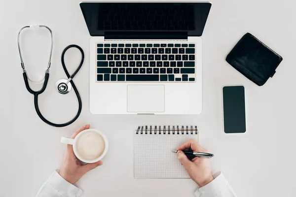Частичное представление врача на рабочем месте с ноутбуком, стетоскопом и чашкой кофе — стоковое фото