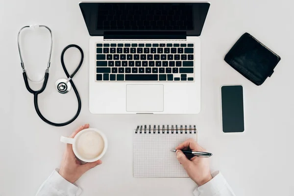 Vista parcial del médico en el lugar de trabajo con computadora portátil, estetoscopio y taza de café - foto de stock