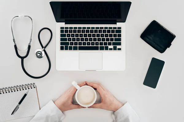 Partial view of doctor at workplace with laptop, stethoscope and cup of coffee — Stock Photo