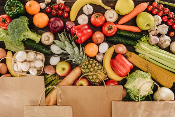 Верхний вид бумажных пакетов, покрывающих овощи и фрукты на деревянном столе, концепция продуктов — стоковое фото