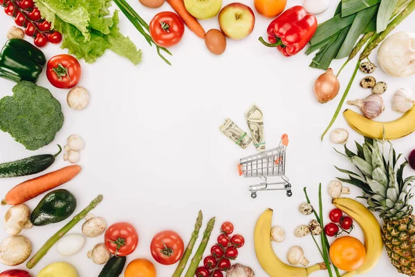 Vista superior de carrinho de compras pequeno com dólares entre legumes e frutas isoladas em branco — Fotografia de Stock