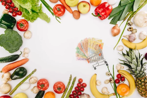 Vista superior del carrito de la compra con dinero en efectivo entre verduras y frutas aisladas en blanco - foto de stock