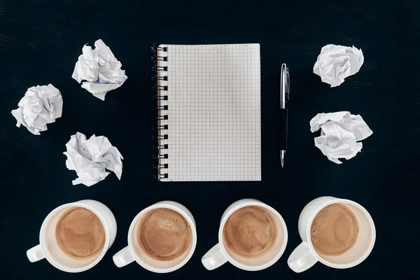 Vista superior del bloc de notas en blanco con papeles arrugados desordenados y tazas de café en fila aisladas en negro - foto de stock