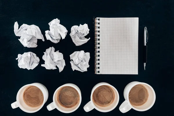 Vista superior de notebook em branco com papéis amassados e xícaras de café em fileira isolado em preto — Fotografia de Stock