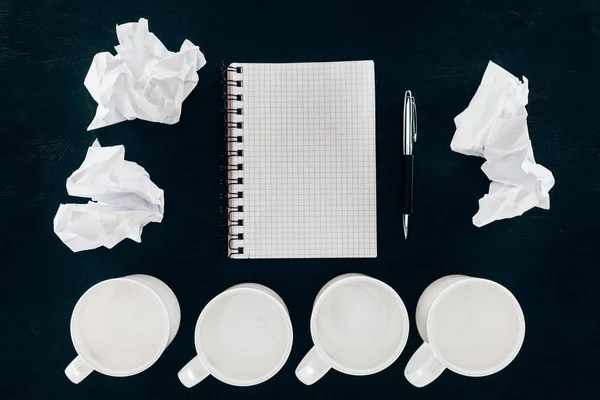 Vue du dessus du bloc-notes vierge avec des papiers froissés et des tasses vides dans la rangée isolée sur noir — Photo de stock