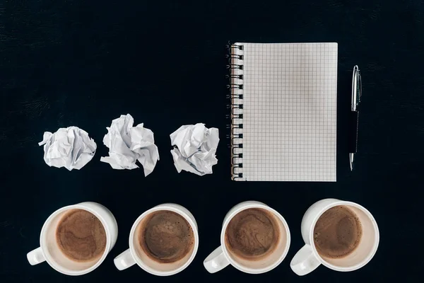 Vista superior do bloco de notas em branco com papéis amassados e xícaras de café em fileira isolado em preto — Fotografia de Stock