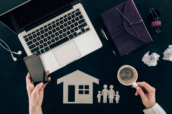 Cropped shot of family lawyer using smartphone at workplace with house and family cut out figures isolated on black — Stock Photo