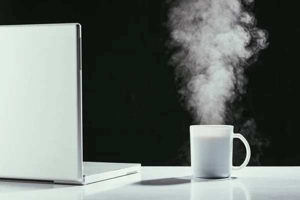 Portatile con tazza di tè fumante sul tavolo isolato su nero — Foto stock