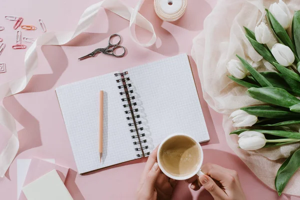 Cropped view of woman holding coffee cup on pink surface with tulips and notepad — Stock Photo