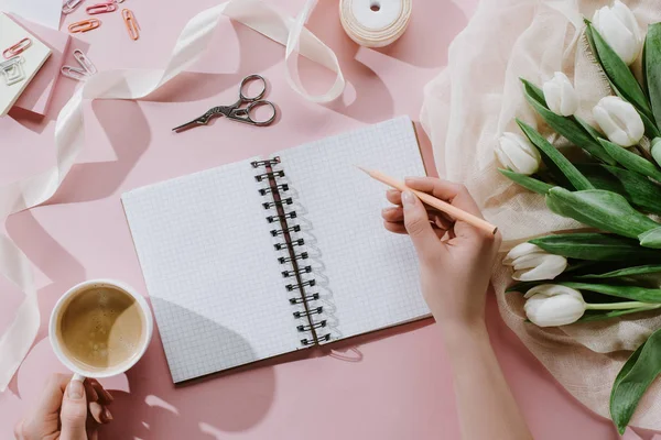 Vista recortada de la mujer escribiendo en bloc de notas en la superficie de color rosa con tulipanes y taza de café - foto de stock