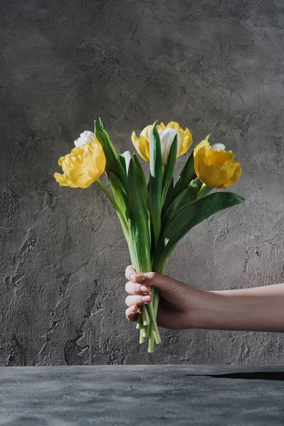Vista recortada de la mujer sosteniendo flores de tulipán amarillo y blanco en la superficie gris - foto de stock