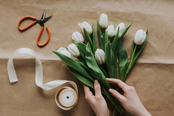 Cropped view of woman holding white tulips on craft paper with ribbon and scissors — Stock Photo
