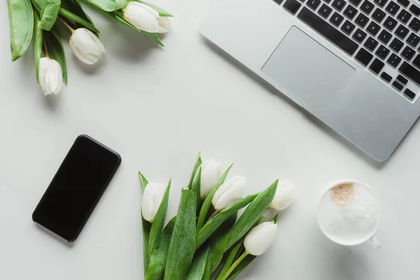 Cama plana con taza de café, portátil, teléfono inteligente y tulipanes blancos en la superficie blanca - foto de stock