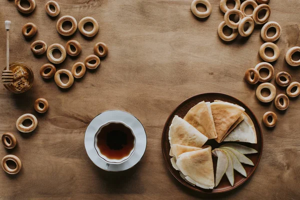 Вид на вкусные домашние блинчики с чашкой чая, бубликами и медом на деревянном столе — стоковое фото
