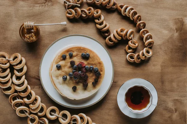 Vista superior de panquecas frescas com bagas, xícara de chá, mel e bagels na mesa de madeira — Fotografia de Stock