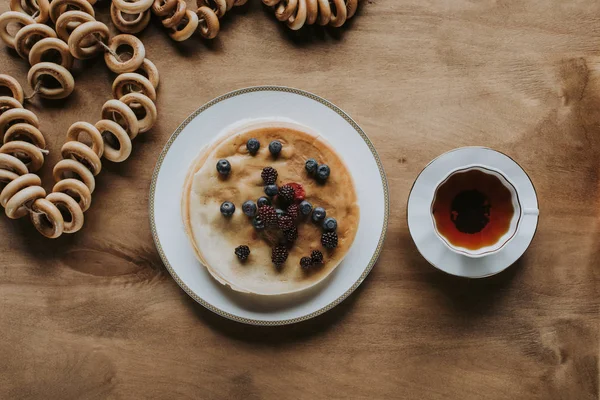 Top view of tasty pancakes with berries, cup of tea and bagels on wooden table — Stock Photo