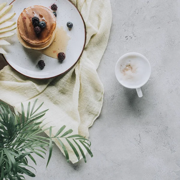 Blick von oben auf leckere hausgemachte Pfannkuchen mit Beeren und Cappuccino auf grau — Stockfoto