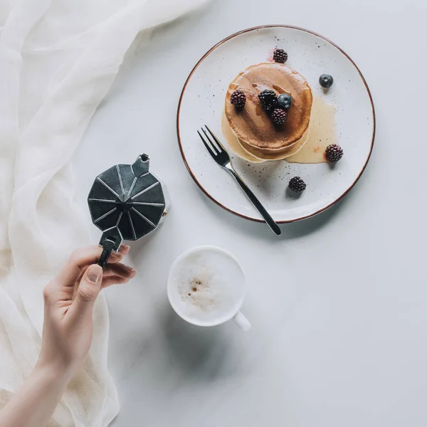 Vista superior de la persona sosteniendo la cafetera mientras come panqueques dulces con bayas para el desayuno - foto de stock