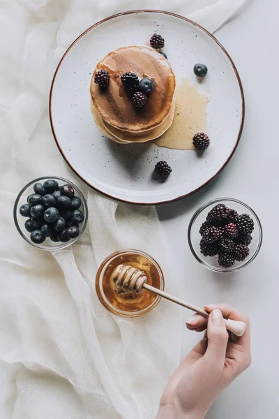 Tiro recortado de la persona comiendo panqueques caseros con bayas frescas y miel en gris - foto de stock