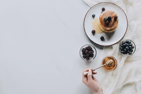 Draufsicht der Person, die frische hausgemachte Pfannkuchen mit Beeren und Honig auf grau isst — Stockfoto