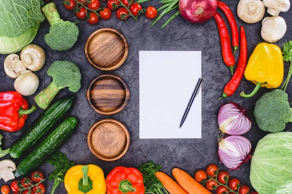 Vista superior de tres platos de madera vacíos, papel en blanco con bolígrafo y verduras frescas surtidas en negro - foto de stock