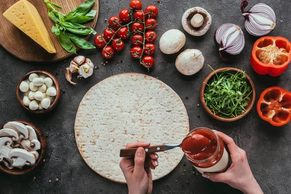 Tiro cortado de mulher espalhando ketchup na massa de pizza crua na mesa de concreto — Fotografia de Stock