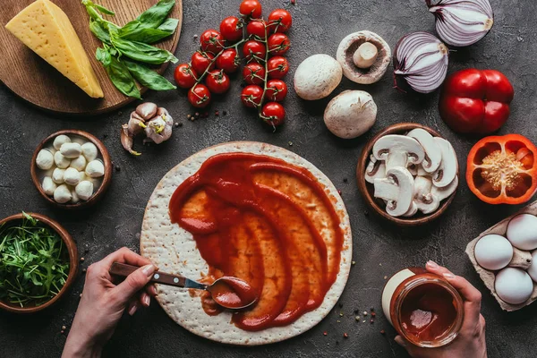 Tiro recortado de la mujer que extiende ketchup sobre la masa de pizza en la mesa de hormigón - foto de stock