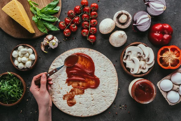 Plan recadré d'une femme étalant du ketchup sur de la pâte à pizza sur une surface en béton — Photo de stock