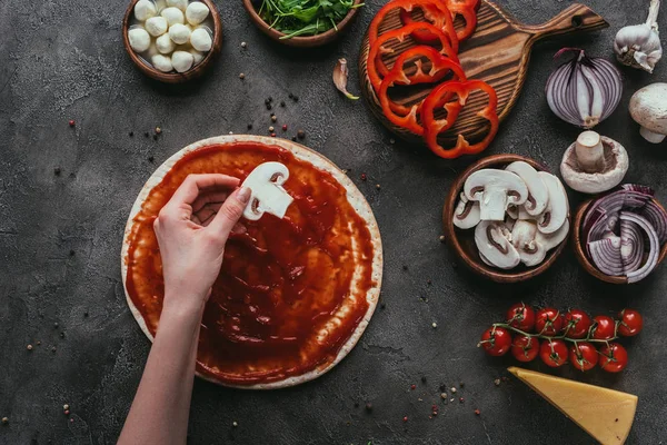 Tiro recortado de la mujer poniendo rodaja de champiñón en masa de pizza en la pizza en la mesa de hormigón - foto de stock