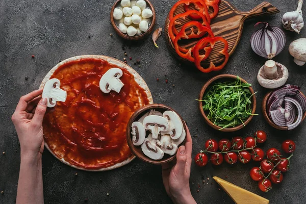 Tiro recortado de la mujer esparciendo rodajas de champiñones sobre la pizza en la mesa de hormigón — Stock Photo