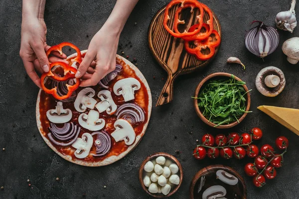 Cropped shot of woman spreading bell pepper slices onto pizza on concrete table — Stock Photo