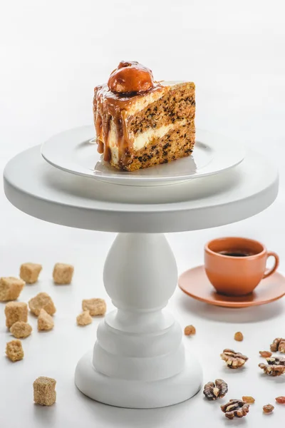 Pedazo de delicioso pastel de caramelo en soporte de pastel blanco, café en la mesa — Stock Photo
