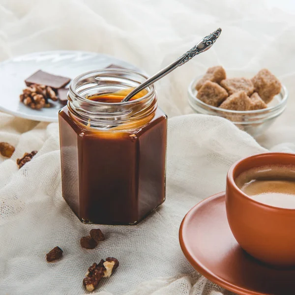 Appetizing jar of caramel jam and cup of coffee on tablecloth — Stock Photo