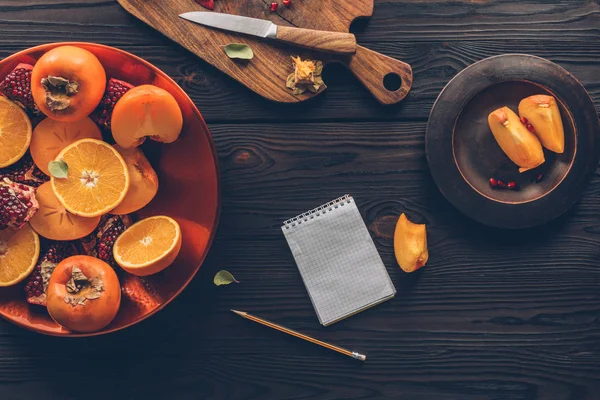 Top view of persimmons, oranges and pomegranates with notebook on table — Stock Photo