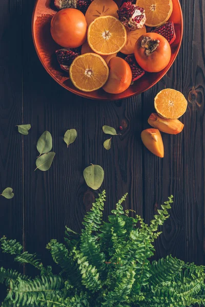 Top view of persimmons with cut oranges and pomegranates on plate and green plant on table — Stock Photo