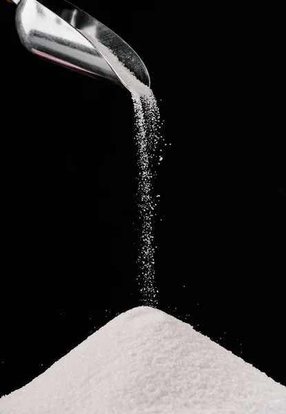 Azúcar que cae de la cucharada de metal en la pila aislada en negro - foto de stock
