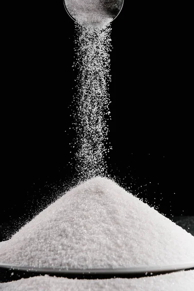 Azúcar que cae de la cucharada de metal en la pila en la placa aislada en negro — Stock Photo