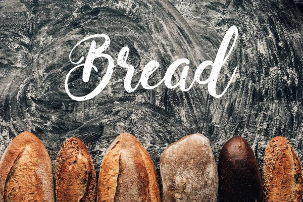 Top view of arranged loafs of bread on dark tabletop with flour and bread lettering — Stock Photo